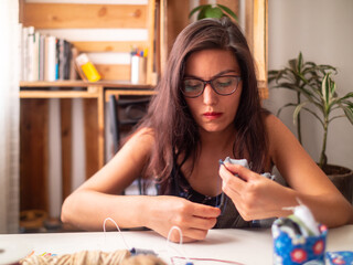 Fototapeta premium Young Attractive Hispanic Woman in Black Dress with Glasses is Quietly Sewing at Home