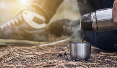 Close up of male hand holding a thermo and pouring a hot beverage in the small mug on the forest floor. Man sits on the ground and pours a tea or coffee from a metal thermos bottle into a cup. - Powered by Adobe