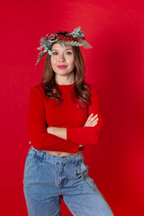 portrait of a smiling young woman in a Christmas wreath, turtleneck and jeans, with her arms crossed on her chest, on a red background