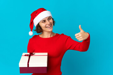Fototapeta na wymiar English girl with christmas hat holding a present isolated on blue background giving a thumbs up gesture