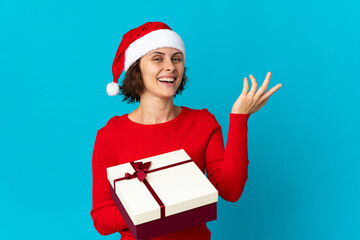 English girl with christmas hat holding a present isolated on blue background extending hands to the side for inviting to come