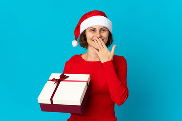 English girl with christmas hat holding a present isolated on blue background happy and smiling covering mouth with hand