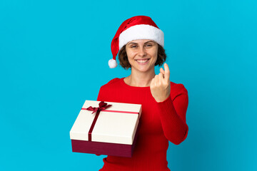 English girl with christmas hat holding a present isolated on blue background doing coming gesture