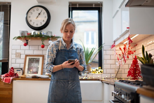 Blond woman wearing blue apron standing in kitchen, using mobile phone.