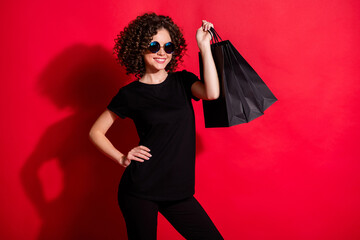 Naklejka premium Photo of cheerful shiny wavy lady dressed casual black outfit eyewear holding shopping bags isolated red color background