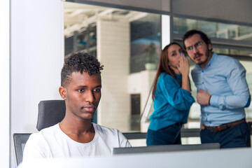 Black man is working on desk and there are two colleagues are gossiping at background behind of him