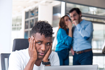 A Sad looking Black man is working on desk and there are two colleagues are gossiping at background...