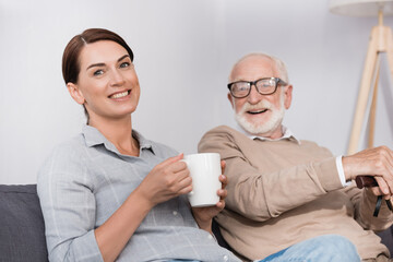 happy woman with cup of tea looking at camera while sitting near cheerful father
