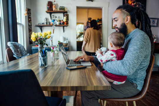 Dad feeding baby while working from home on laptop computer at dining room table