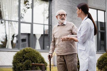 geriatric nurse supporting aged man strolling with walking stick