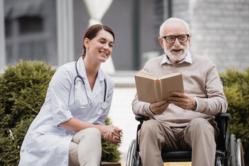 cheerful geriatric nurse near aged disabled man reading book in wheelchair outdoors