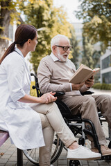 Plakat geriatric nurse sitting near aged disabled man reading book outdoors