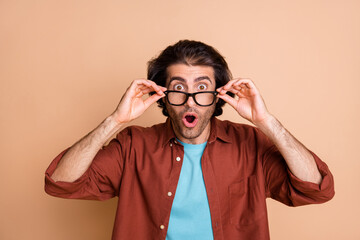Photo of amazed young guy arms hold eyewear open mouth wear glasses brown t-shirt isolated beige color background