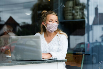 Woman wearing face mask sitting alone at a cafe table with a laptop computer, looking out