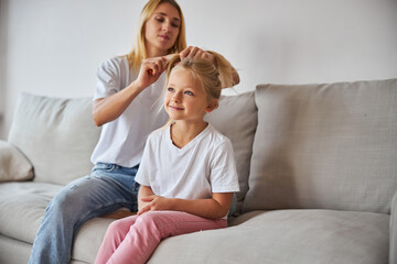 Young Caucasian woman doing girl hair while sitting on the sofa in living room