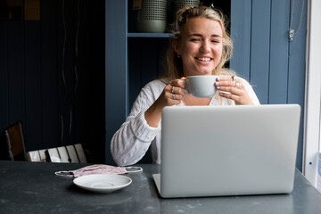 Young bwoman sitting alone at a cafe table with a laptop computer, drinking coffee