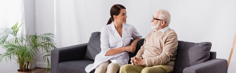 smiling geriatric nurse talking to aged patient while sitting on sofa, banner