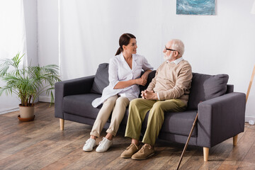 Obraz premium brunette social worker and aged man talking while sitting on sofa at home