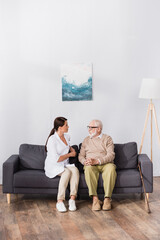 aged man in eyeglasses talking to geriatric nurse while sitting on sofa at home