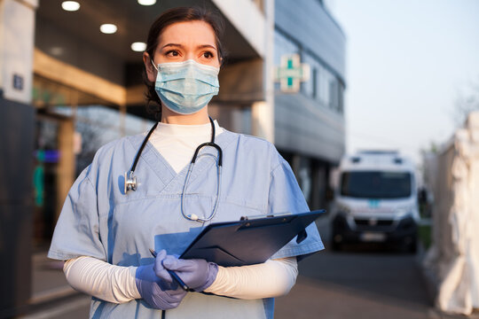 Young female EMS key worker doctor in front of healthcare ICU facility,wearing protective PPE face mask equipment,holding medical lab patient health check form,UK&US COVID-19 pandemic outbreak crisis 