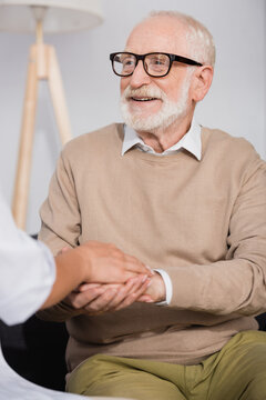 smiling senior man holding hands with social nurse at home on blurred foreground