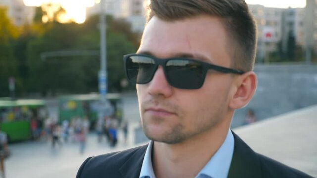 Portrait of young businessman in sunglasses walking in city street. Handsome business man stepping at town square and looking at the camera. Face of confident guy commuting to work. Close up