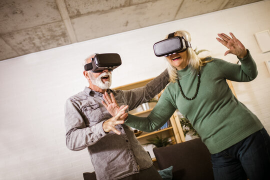 Senior couple play virtual reality glasses headset playing video game and have fun together in living room at home.