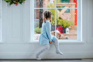 Girl in winter dress sitting near the window with Christmas background. Snowing.