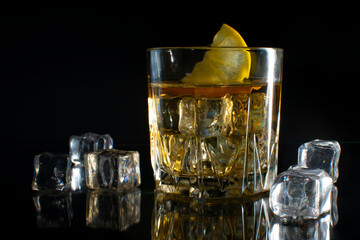 A glass of whiskey with ice with a slice of lemon on a black table with reflection. On a black background.
