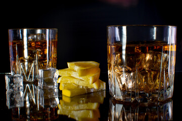 Two glasses with whiskey on ice with sliced lemon on a black table with reflection. On a black background.