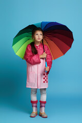 little child girl with multicolored umbrella in pink rain coat and rubber boots looking up on blue background.