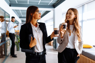 Young business women discussing something and drinking coffee while walking down the office...