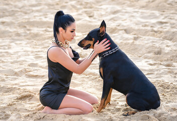Attractive woman in a black dress with a doberman sitting on the sand opposite each other.