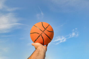 The hand of a male athlete holds a basketball ball against a blue cloudy sky. Sports activities on the playground.