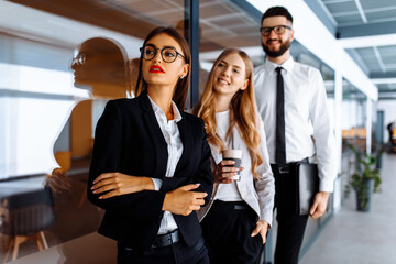 Group of attractive business people standing and communicating together, holding cups, in a modern office. Coffee break.