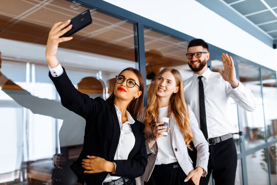 Group of attractive business people standing and chatting together holding coffee cups and taking selfie on mobile phone office