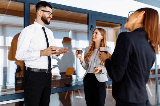 Group of attractive business people standing and communicating together, holding cups, in a modern office. Coffee break.
