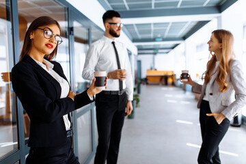 Young business people standing and chatting together, holding cups, in a modern office. Coffee break.