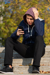 Relaxed teenage boy sitting on the steps outdoors using mobile phone and chatting with friends online. Contented caucasian teenager in hood looking at smartphone screen.