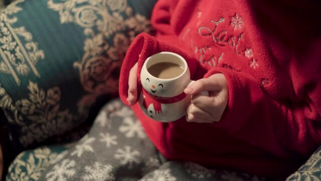 girl in Christmas pajamas drinking coffee in a cup shaped like a snowman. teenager on blue sofa warming his hands with a hot drink. hot milk in cup and hands, woman with tea.
