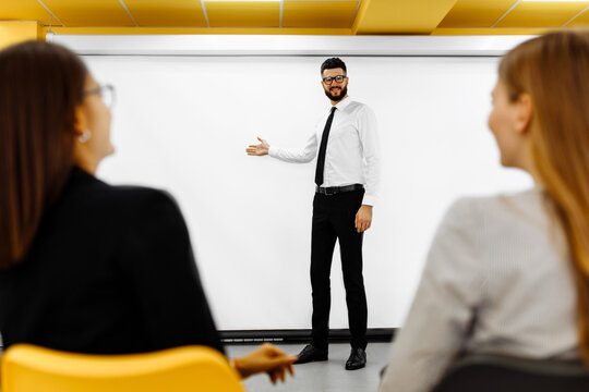 Handsome young businessman making presentation to his colleagues and pointing at white background in conference room at office