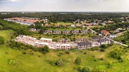 Aerial view on estate of multi-family houses under construction. House In basic state. Wooden roof structure under construction. House located on green plot in small village. Green meadows around.