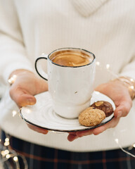 A girl dressed in a white sweater holds a white cup of coffee in her hands. There are cookies on the saucer. Warming Christmas drink. Christmas coffee. Coziness in Christmas. Hot coffee in hands.