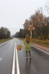 A girl in a green dress with autumn leaves in her hands is walking along the highway