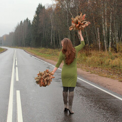 A girl in a green dress with autumn leaves in her hands is walking along the highway