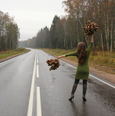 A girl in a green dress with autumn leaves in her hands is walking along the highway