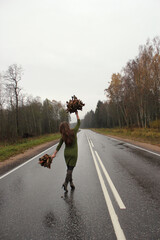 A girl in a green dress with autumn leaves in her hands is walking along the highway