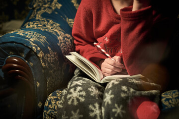 girl in red and gray Christmas pajamas with snowflakes on sofa. teenager writing in notebook with Christmas pencil. pencil in the shape of a caramel stick and written message