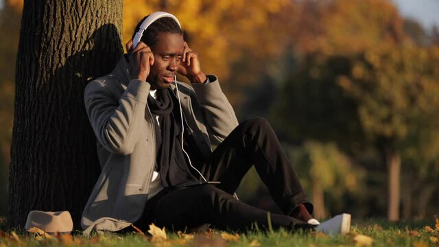 African man listening to music in headphones leaning on a tree trunk at the fall day