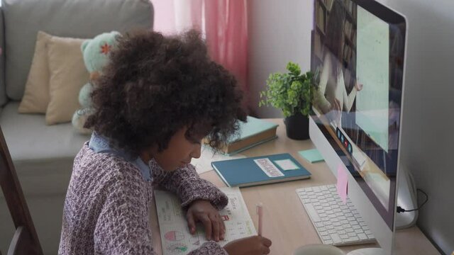 Smart small african school child girl online learning with online remote teacher or web tutor studying at home on computer taking notes. Tutoring, homeschool social distance virtual education concept.